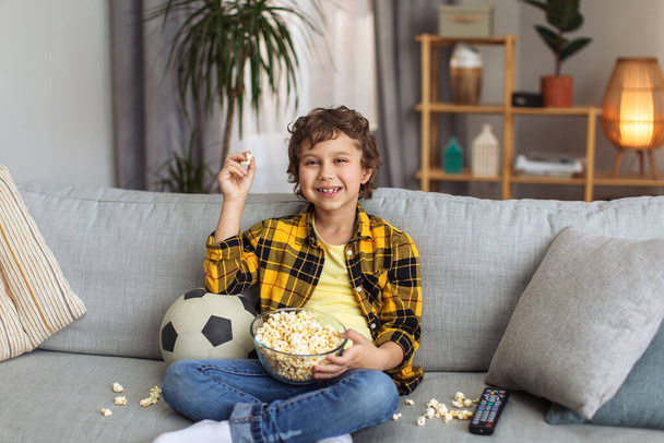 Home alone. Happy little boy having fun, watching football on tv and eating popcorn, sitting on messy sofa and laughing, free space - Foto, Bild