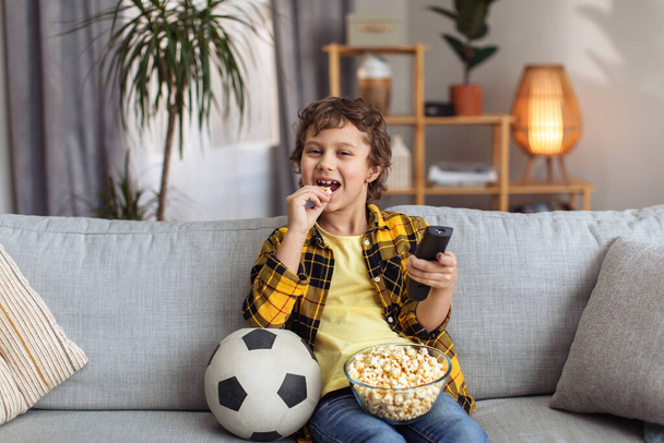 TV entertainment. Happy little boy spending weekend at home, watching sport match and eating popcorn, resting on sofa in evening, free space - Fotó, kép