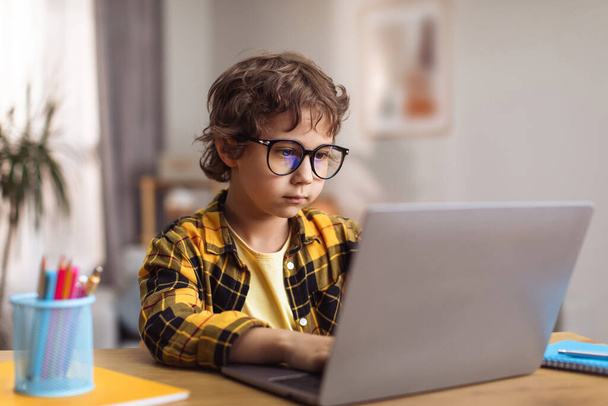 Smart generation. Concentrated little boy wearing eyeglasses typing on laptop at home, creating new game online, sitting at desk, free space - Foto, afbeelding