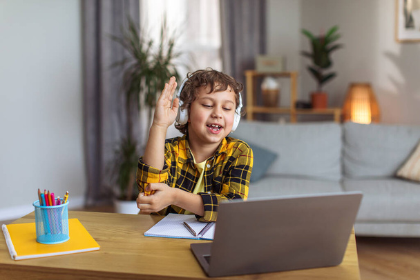 Online education. Clever little boy raising hand, want to answer question, studying distantly from home via laptop app, free space - Foto, afbeelding