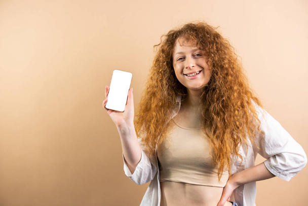 Redhead woman holding newest smartphone with empty screen, enjoying new application for mobile phone over golden studio background, creative image. High quality photo - Фото, зображення
