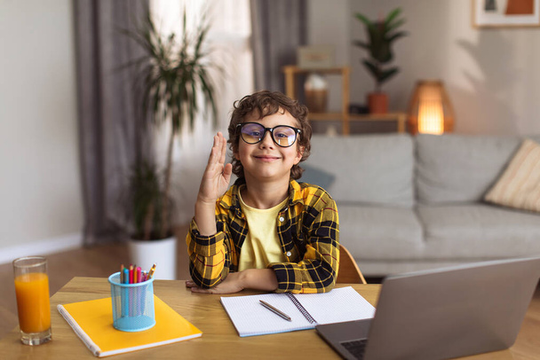 E-learning. Cute schoolboy in eyeglasses raising hand and smiling to camera, studying distantly at home interior, free space - Fotó, kép