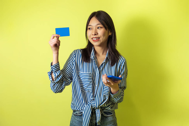 Мобільні покупки. Щаслива японська леді тримає кредитну картку та смартфон, роблячи платежі онлайн стоячи на жовтому тлі в студії. Концепція додатків Інтернет банкінгу
 - Фото, зображення