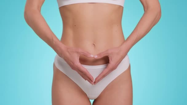 Female health concept. Close up shot of slim unrecognizable woman in underwear showing heart gesture with her hands near belly bottom, blue studio background, slow motion - Video