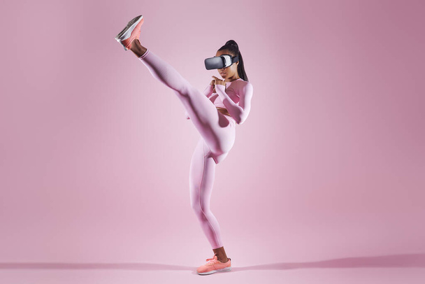 Woman in virtual reality headset practicing in kicking while standing against pink background - Foto, imagen