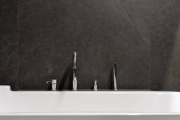 closeup of detail interior in modern bathroom in hotel with white bath, silver faucet and shower and dark grey tile on wall indoors - Foto, imagen