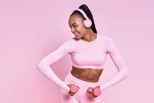 Attractive African woman in headphones exercising with dumbbells against pink background - Foto, imagen
