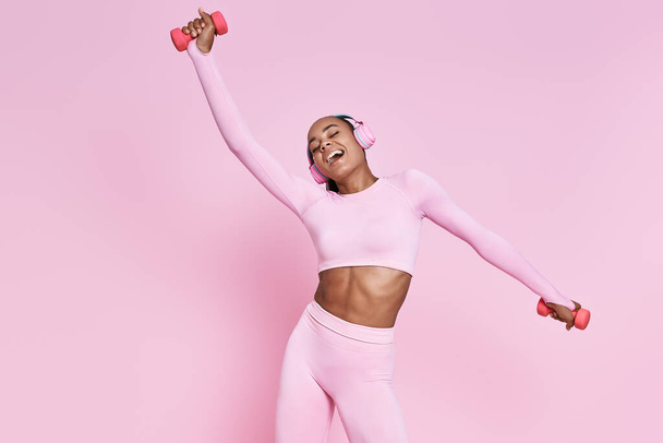 Playful African woman in headphones exercising with dumbbells against pink background - Foto, afbeelding