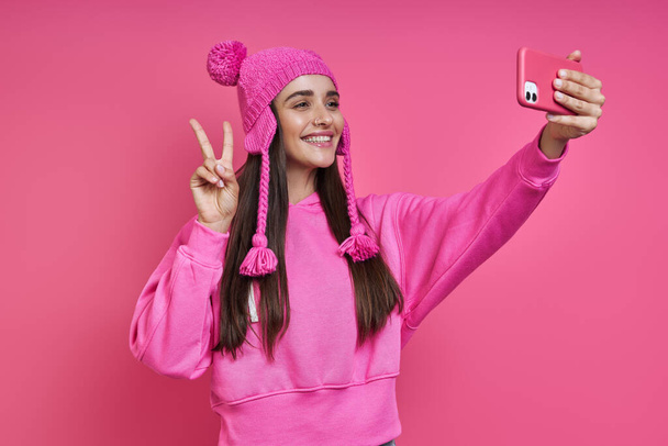 Παιχνιδιάρα νεαρή γυναίκα σε funky καπέλο κάνει selfie και gesturing σε ροζ φόντο - Φωτογραφία, εικόνα