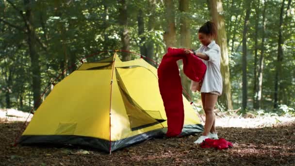 Размещение договоренности. Молодая позитивная африканская американка кладет спальный мешок в палатку, готовится к ночевке в лесу, замедленное движение - Кадры, видео