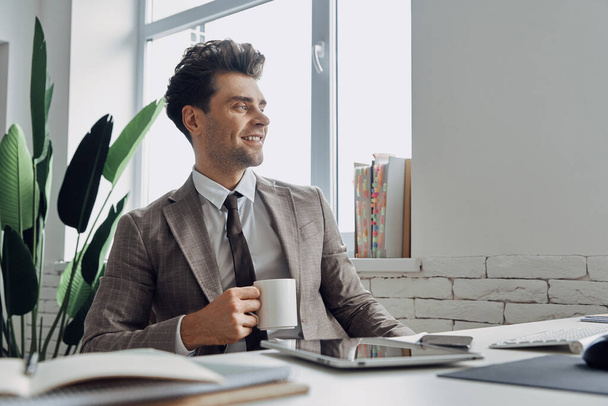 Handsome man holding coffee cup and smiling while sitting at his working place in office - Photo, Image