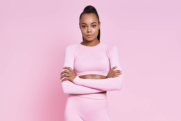 Confident young African woman in sports clothing standing against pink background - Zdjęcie, obraz