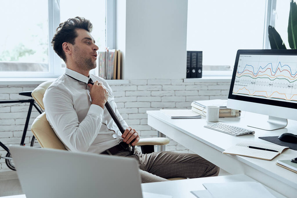 Relaxed young man adjusting his necktie while sitting at his working place in office - Foto, Bild