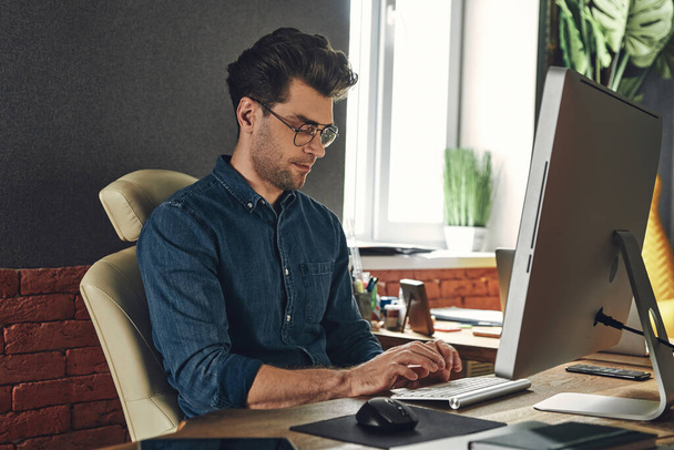 Handsome young man using computer while sitting at his working place in office - Photo, Image