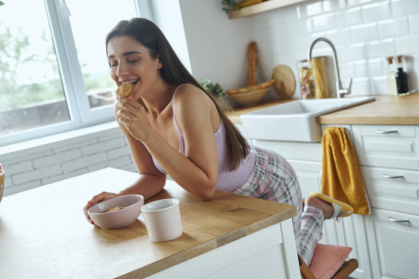 Beautiful young woman eating cookies while sitting at the kitchen counter - Foto, afbeelding