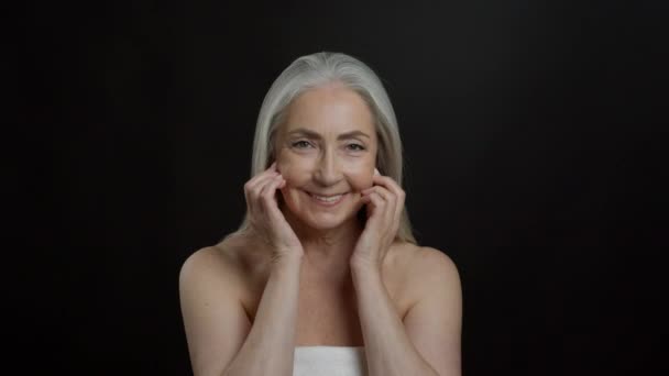 Face yoga concept. Pretty senior grey haired woman with bare shoulders touching her face, doing light massage, black studio background, slow motion - Footage, Video