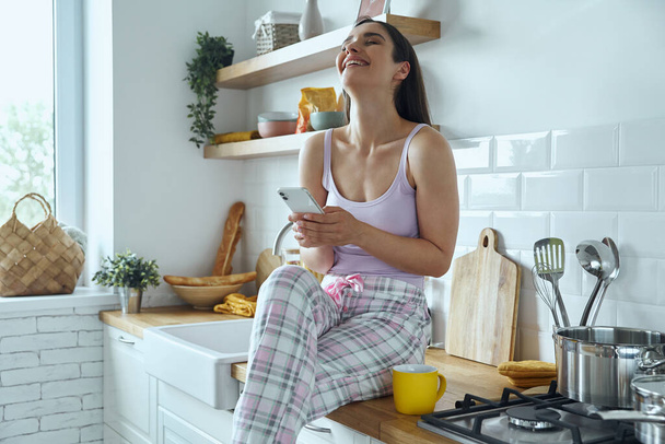 Joyful young woman holding smart phone and smiling while sitting on the kitchen counter - Foto, afbeelding