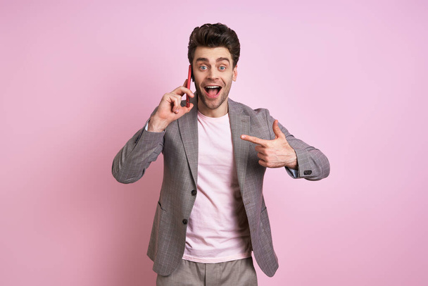Excited man in suit talking on mobile phone and pointing it while standing against pink background - Foto, immagini