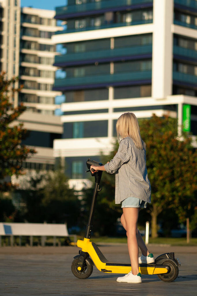 Young beautiful slender woman in denim shorts, shirt, riding an electric scooter in the park in summer, photo from the back.Modern girl,new generation, electric transport, ecological transport, sunset - Foto, Bild