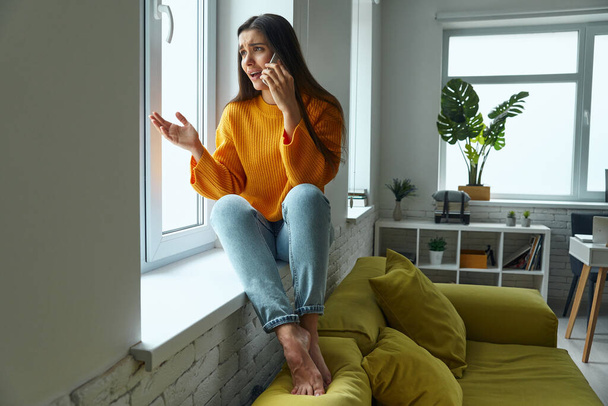 Worried woman talking on mobile phone and gesturing while sitting on the window sill - Photo, image