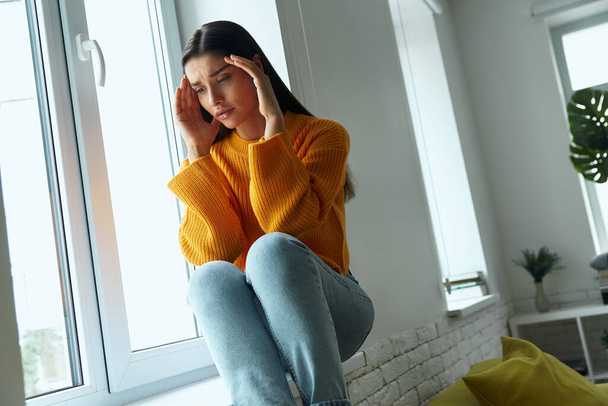 Depressed young woman touching head with hands while sitting on the window sill - Foto, Bild