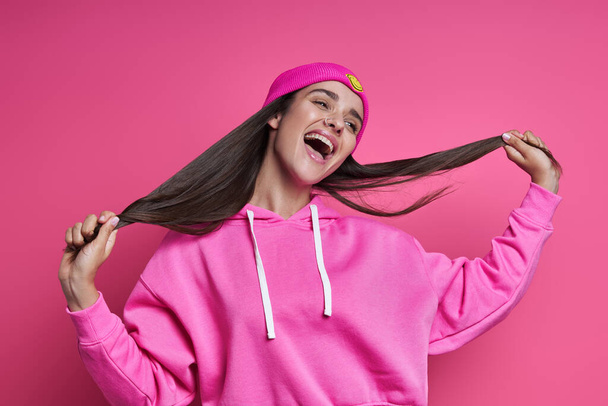 Excited young woman in hooded shirt holding her long hair against pink background - Фото, зображення