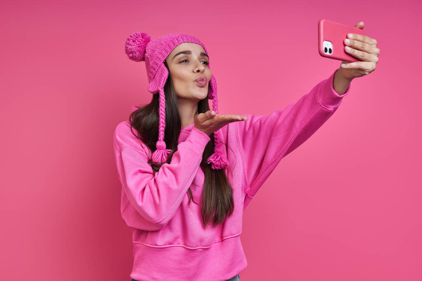 Παιχνιδιάρα νεαρή γυναίκα σε funky καπέλο κάνει selfie και φυσάει ένα φιλί σε ροζ φόντο - Φωτογραφία, εικόνα