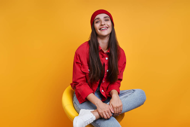 Beautiful young woman smiling while sitting on the chair against yellow background - Foto, afbeelding
