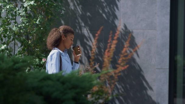 Attraente ragazza che beve caffè passeggiando quartiere del centro sulla mattina di sole. Carefree manager donna d'affari pendolare al lavoro tenere bevanda tazza. Sorridente donna afroamericana andare strada con borsa a tracolla - Foto, immagini