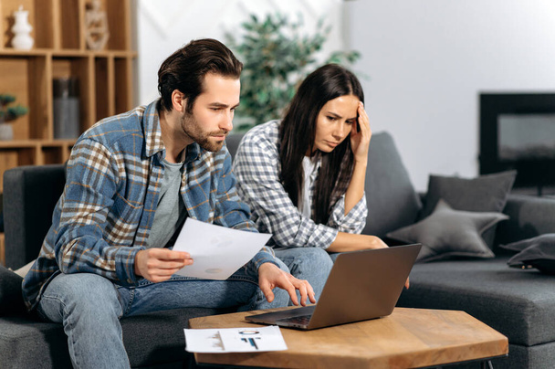 Frustrated caucasian couple sitting on the couch, the guy and the girl are freelancers, work from home, use a laptop, look at graphs, are unhappy with the results, low profit, thinking about strategy - 写真・画像