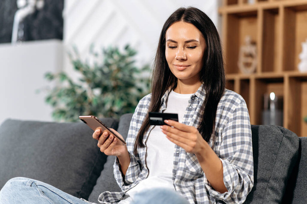 Online shopping. Satisfied caucasian woman makes purchases on the Internet choosing goods in online stores, sits on a sofa in the living room, holds a smartphone and a bank card, pays for goods, smile - Fotografie, Obrázek