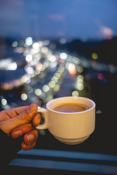 Hand holding a hot latte coffee in a white cup over a blurry background of colorful morning city lights - Foto, afbeelding