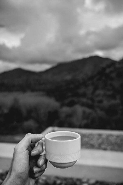 Hand holding a hot latte coffee in a white cup over a blurry background of a landscape with forest, mountains, cloudy sky and a terrace with stones (in black and white) - Foto, Imagen
