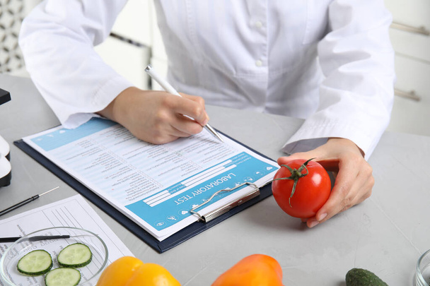 Scientist with laboratory test form and fresh tomato at table indoors, closeup. Poison detection - Фото, изображение