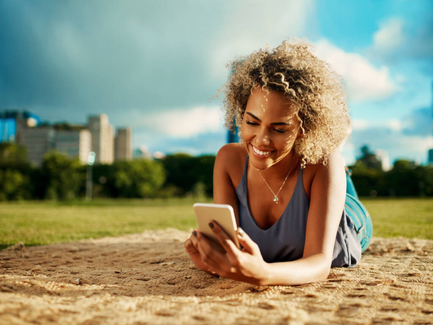 Texting a special someone... an attractive young woman using a smartphone while lying down at a park - Foto, Imagen