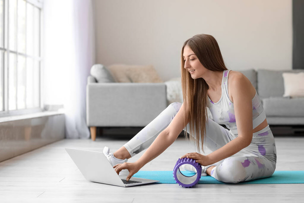 Young woman with foam roller using laptop at home - Fotoğraf, Görsel