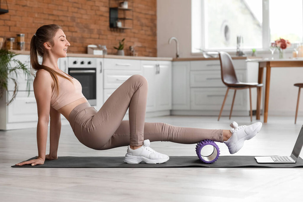 Young woman training with foam roller and laptop in kitchen - Zdjęcie, obraz