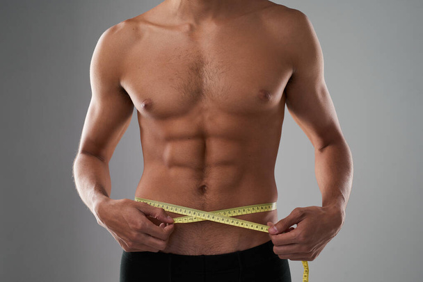 Its all about the measurements for him. Studio shot of an unrecognizable shirtless man measuring his midsection against a grey background - Foto, imagen