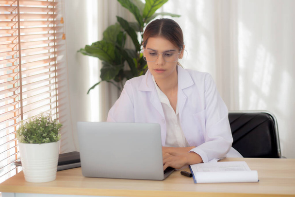 Young woman doctor using laptop computer and looking document paper about patient for diagnostic at clinic, physician female working and reading document on clipboard, one person, medical concept. - 写真・画像