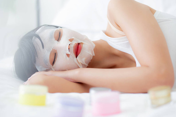 Beautiful young asian woman with sheet facial mask on bed at bedroom, beauty girl lying applying cosmetic and makeup with skin care face for wrinkle at home, one person, skincare treatment and health. - Photo, Image