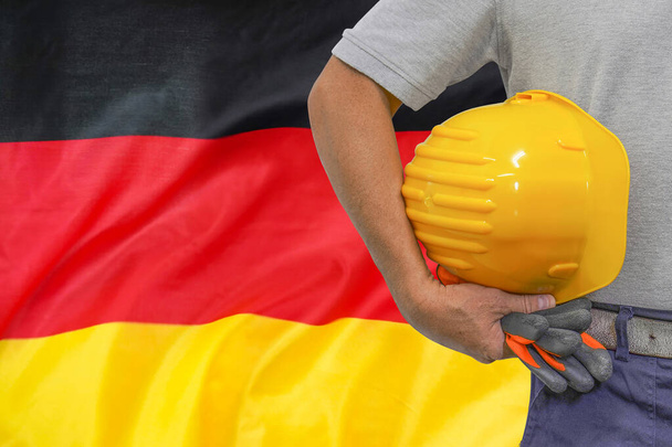 Κοντινό πλάνο του hard hat holding από τον εργαζόμενο οικοδομών στο φόντο σημαία της Γερμανίας. Χέρι εργάτη με κίτρινο σκληρό καπέλο και γάντια. Έννοια των εργαζομένων στη βιομηχανία, τις κατασκευές και τη βιομηχανία στη Γερμανία - Φωτογραφία, εικόνα