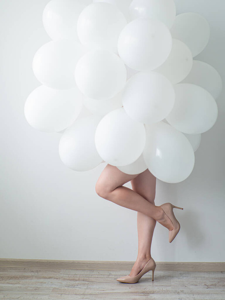 Woman in beige high heels holding white color balloons on a white wall background. Legs and a lot of white air balloons. Happy birthday anniversary. White decoration. Copy space - Fotografie, Obrázek