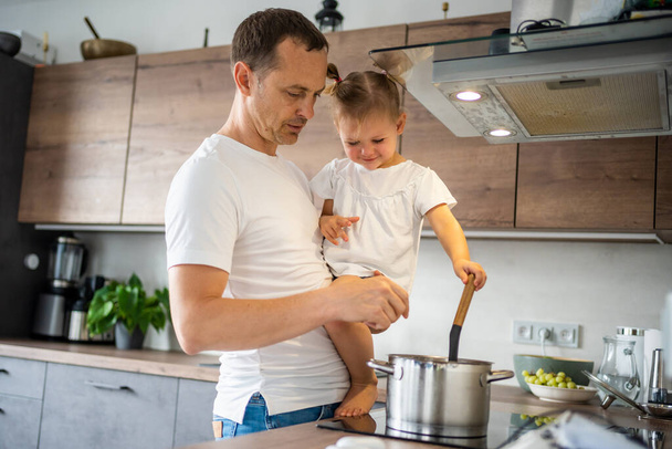 Father with his daughter on his hands cooking soup together in modern home kitchen. High quality photo - Zdjęcie, obraz