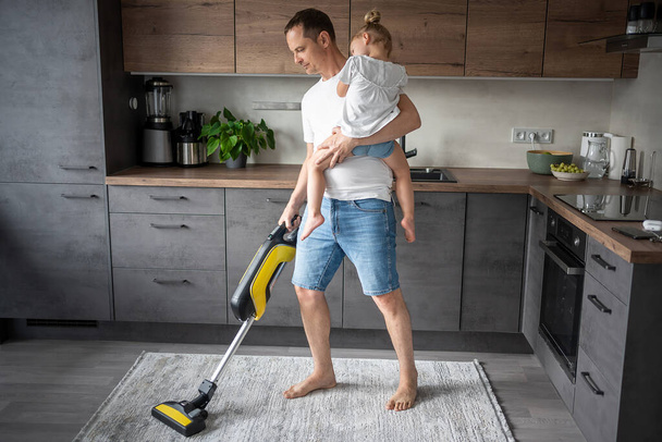 Father with his daughter on his hands vaccuming the floor in the modern home kitchen. High quality photo - Fotografie, Obrázek