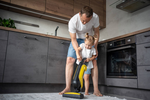 Father and daughter having fun with vacuum cleaner while cleaning at home kitchen. High quality photo - Foto, afbeelding