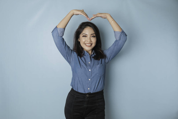 An attractive young Asian woman wearing a blue shirt feels happy and a romantic shapes heart gesture expresses tender feelings - Valokuva, kuva