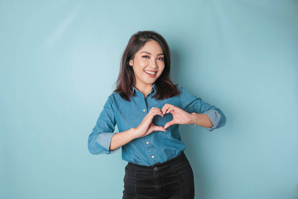 An attractive young Asian woman wearing a blue shirt feels happy and a romantic shapes heart gesture expresses tender feelings - Zdjęcie, obraz