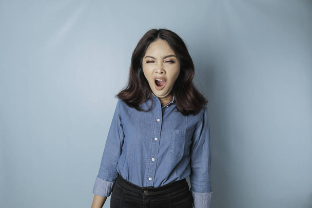 Portrait of sleepy attractive Asian woman wearing a blue shirt, feeling tired after night without sleep, yawning, covering opened mouth with palm - Foto, immagini