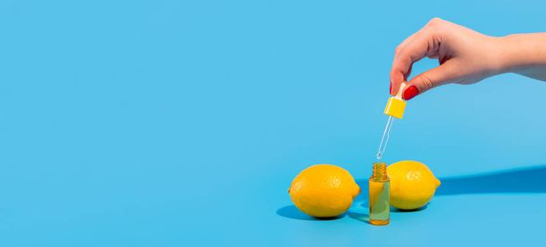 Serum in a transparent bottle with a pipette hold hand with yellow lemons on blue background. Cosmetic moisturizer with retinol in a glass container. Natural beauty organic moisturizing oil. vitamin c - Photo, Image