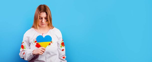Rapariga ucraniana triste em uma camisa bordada com a bandeira da Ucrânia em seu rosto tem um coração de uma bandeira amarelo-azul. O conceito de participação do povo ucraniano na guerra com a Rússia - Foto, Imagem